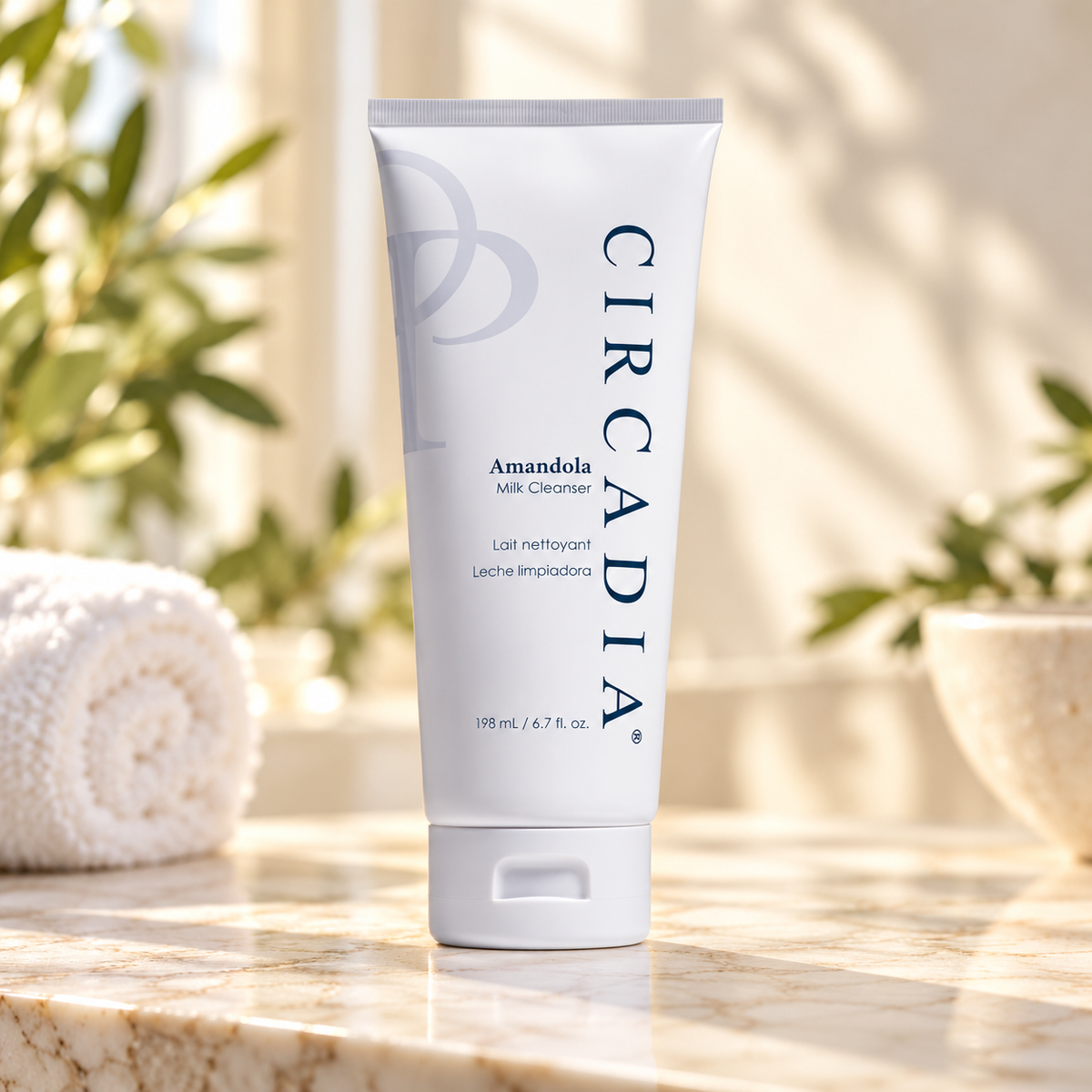 A white squeeze tube of Circadia Amandola Milk Cleanser stands on a marble surface in a bright, sunlit bathroom setting with a rolled towel and greenery in the background.