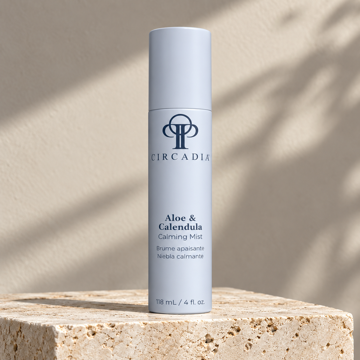 A light blue cylindrical bottle of Circadia Aloe & Calendula Calming Mist stands on a textured stone pedestal against a warm beige wall with soft leaf shadows.