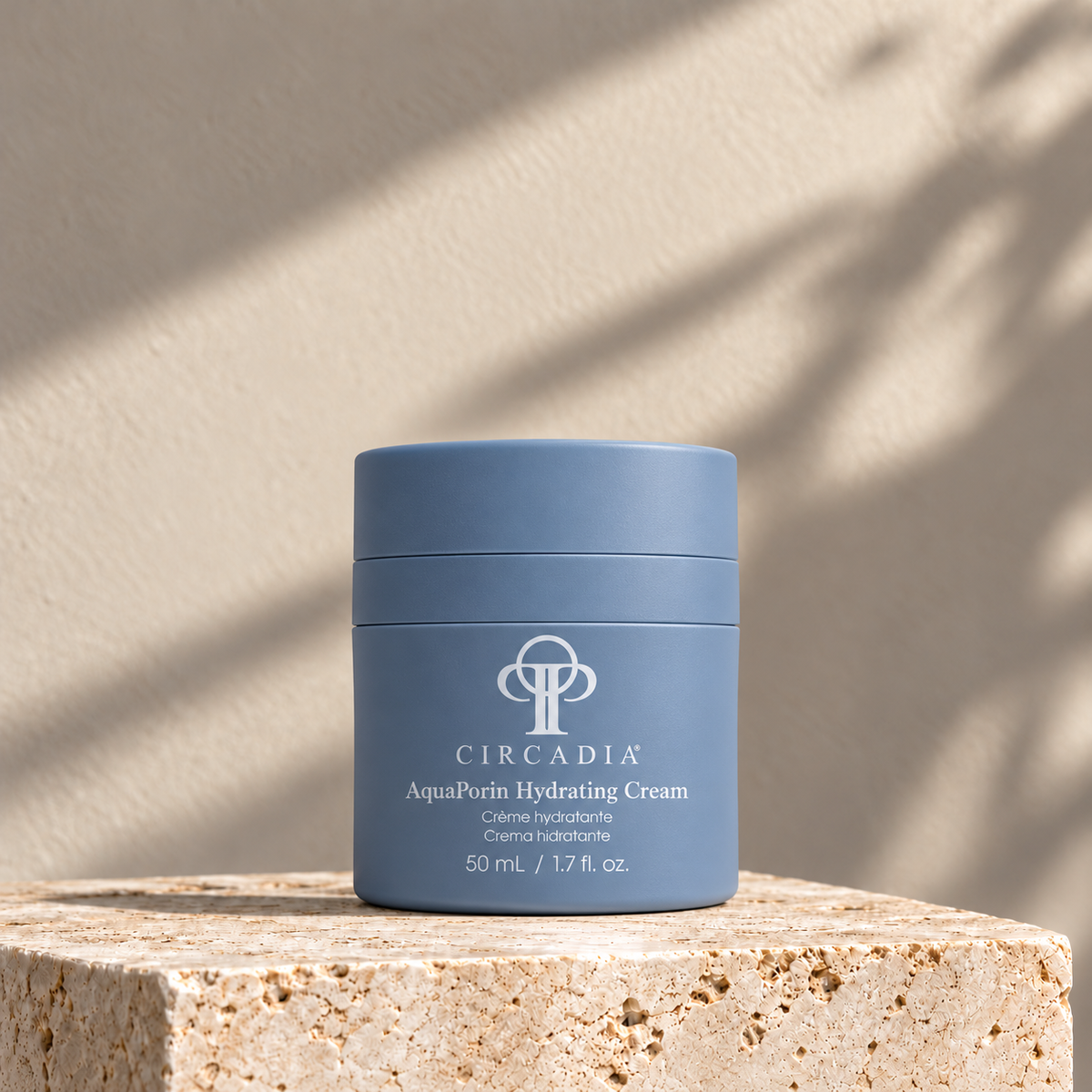 A blue cosmetic jar of Circadia AquaPurin Hydrating Cream sits on a textured stone pedestal against a warm beige wall with soft leaf shadows cast across it.
