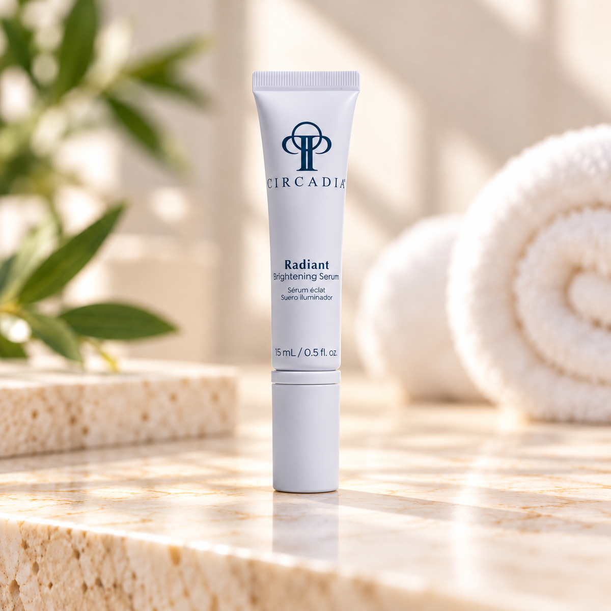 A white tube of Circadia Radiant Brightening Serum stands on a light stone surface with a softly blurred bathroom-like setting, including green leaves and a rolled towel in the background.
