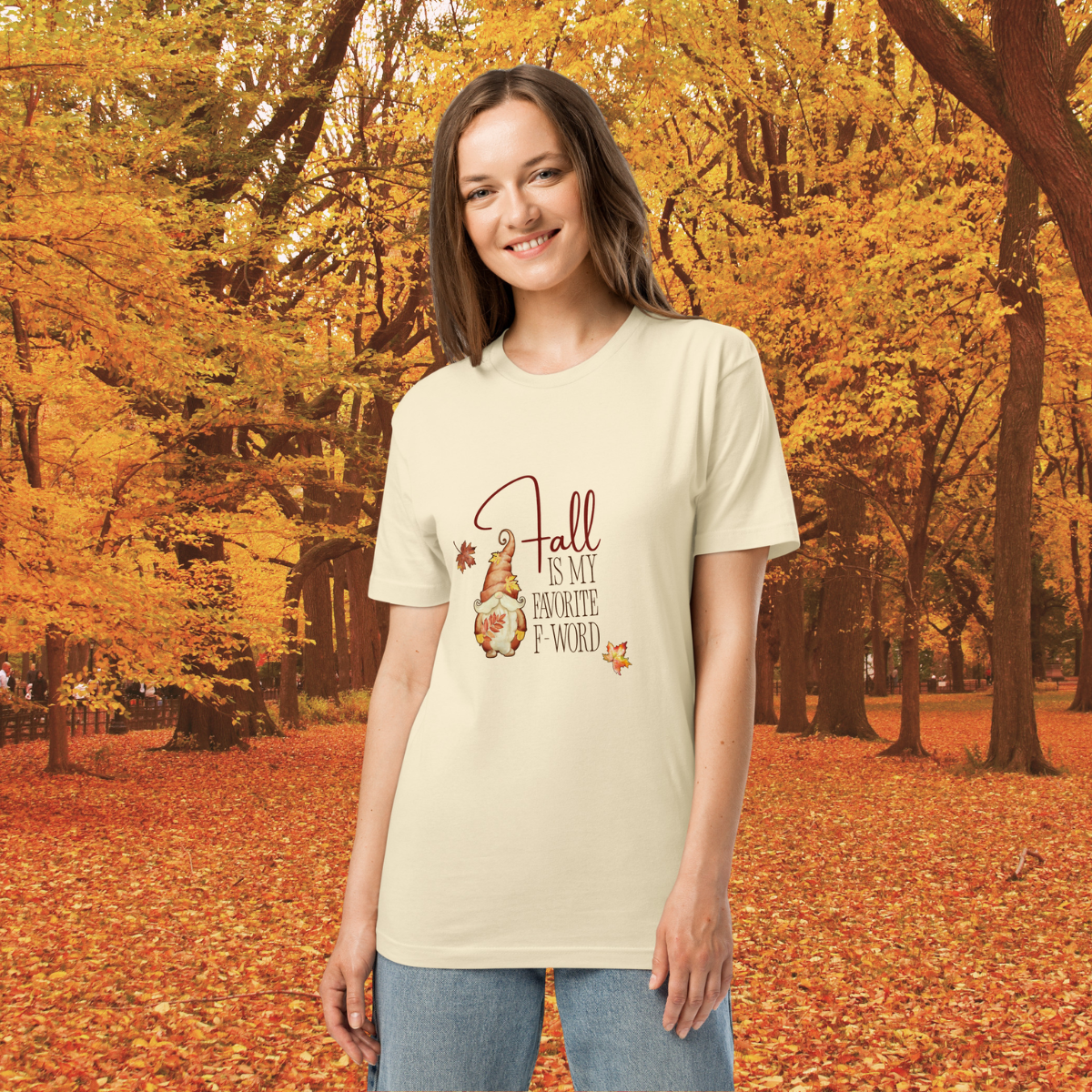 A woman wearing a light beige graphic t-shirt with a fall-themed design that reads 'Fall is my favorite word' alongside a small pumpkin illustration, set against a background of a forest with orange autumn trees and fallen leaves.