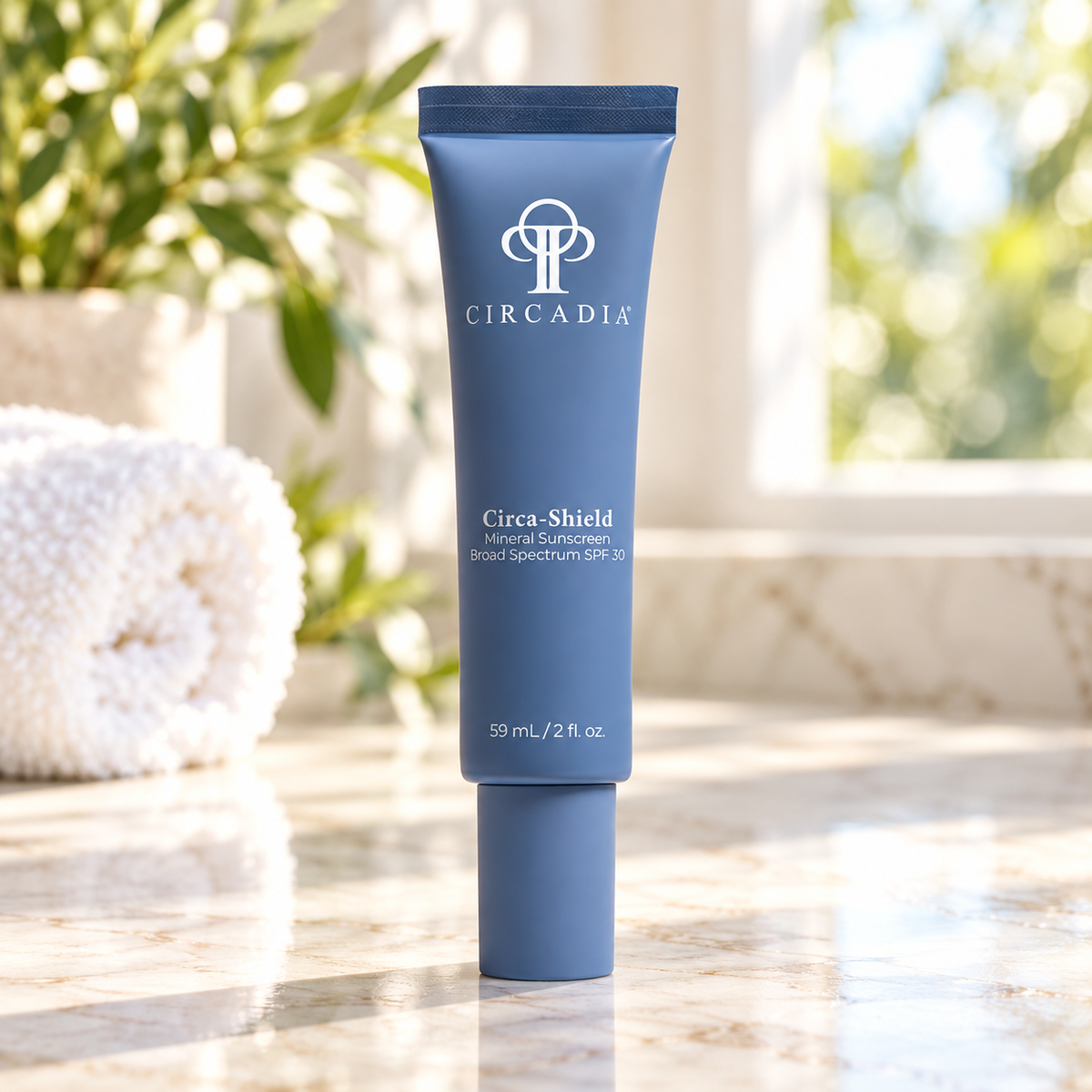 A blue tube of Circadia Circa-Shield Natural Coloration Brast Spectrum SPF 30 sunscreen, standing on a marble countertop with a blurred background of a window, greenery, and a white towel.